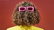 Person with curly hair and their back turned to the camera. They are wearing pink sunglasses on the back of their head, covering the back of the head, not their eyes.
